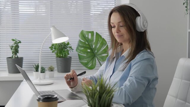 A woman, focused on remote work, wears stylish headphones while typing on her laptop. Vibrant houseplants surround her, enhanced by warm natural light streaming through the window