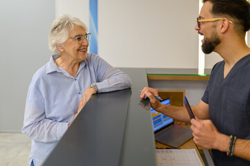 Elderly woman warmly interacting with young doctor at reception counter during hospital visit for medical guidance