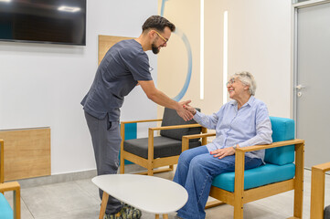 Obraz premium Young male doctor smiling and welcoming elderly woman with handshake during visit in hospital lobby