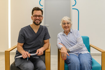 Portrait of confident doctor and patient sitting on chairs and smiling at camera during medical examination at clinic