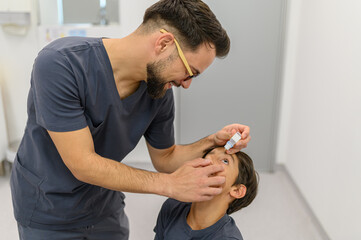 Smiling young male eye doctor putting drops in boy's eye during medical examination room at clinic