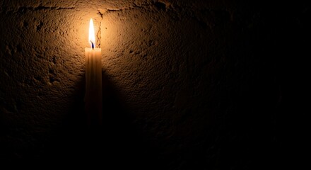 A single lit candle casts a warm glow against a dark, textured wall.