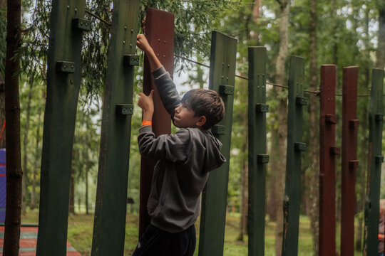 A focused boy in a hoodie climbs vertical wooden poles in an outdoor forest obstacle course, holding on tightly with both hands in a natural forest environment. 