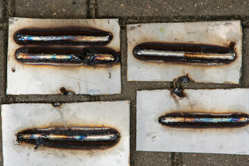 Close-up of several welding test beads on metal plates, showing different welding techniques and heat discoloration. Industrial background with practice welds on steel sheets placed on the ground."