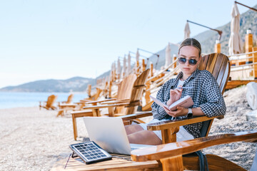 Beach workation digital nomad plans tasks with laptop and notebook on lounge chairs, powered by a...