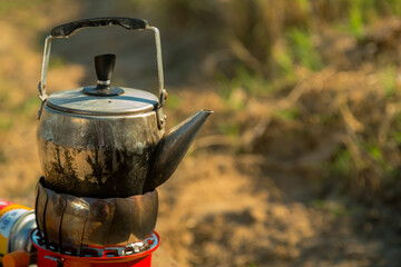 A small metal kettle is boiling on a camping gas burner in nature