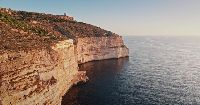 Malta: Golden sunset light reflecting on Dingli rocks cliffs, the highest point of Malta, with a radar dome on top and the Mediterranean Sea calm waters shining below, aerial drone flight