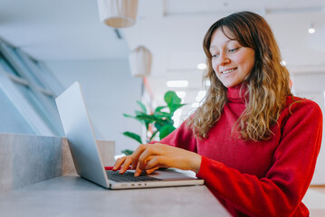 Smiling young woman sitting at a table in a bright, cozy room, using a laptop to search for gifts...