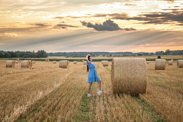 Portrait of a young beautiful dark-haired girl in a summer dress in a village field among bales of...