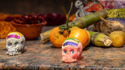 Two skulls are on a table with oranges and other fruits. The skulls are decorated with colorful ribbons. Day of the Dead celebration in Mexico, papel picado crafts, offerings, La Catrina