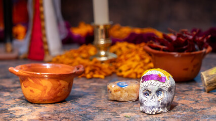 A skull is sitting on a table next to a bowl and a candle. The skull is decorated with a purple band. Day of the Dead celebration in Mexico, papel picado crafts, offerings, La Catrina