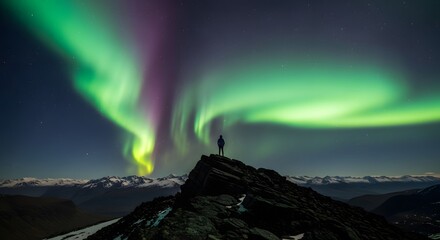 A lone hiker stands on a rocky peak, silhouetted against a vibrant green and purple aurora borealis dancing in the night sky above snowcapped mountains