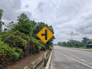 Road traffic sign showing a T junction ahead, standing on roadside with cloudy sky