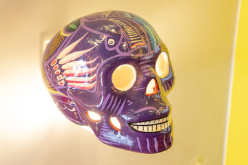 A purple skull with a smiley face on it. The skull is decorated with a colorful design and has a light shining on it. Day of the Dead celebration in Mexico, papel picado crafts, offerings, La Catrina