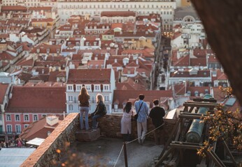 At golden hour, several individuals gaze across a sprawling cityscape of red-tiled roofs from the ramparts of Castelo de Sao Jorge, where an old cannon sits ready for firing