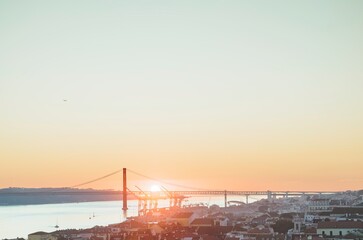Obraz premium The sun sets behind the 25 de Abril Bridge in Lisbon, casting a warm golden glow over the Tagus river and the cityscape. Gentle light fills the sky and reflects on the water