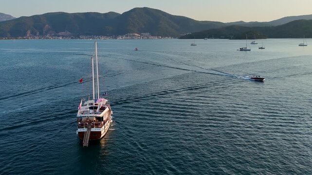 Cinematic Aerial View of a Two-Masted Sailboat Navigating Wide Open Green Sea Water with Beautiful Misty Mountains in the Background. Peaceful Travel Scene at Twilight.