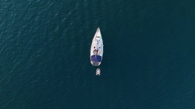 A top down drone view of a white gulet style sailboat with solar panels on its deck anchored in the tranquil blue sea with a small dinghy boat nearby a luxury travel video