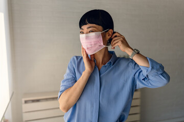 Asian woman in blue shirt smiling gently while adjusting a pink protective mask indoors near a window.