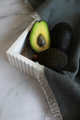 Fresh whole and halved avocados in a white tray with a gray linen cloth on a marble surface.