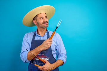 Chilean chef holding barbecue fork looking up, wearing straw hat and apron, on blue background,...