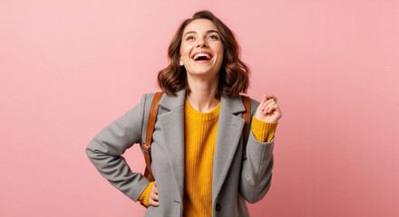 A happy young woman laughing with joy. Stylish student in a yellow sweater and grey coat posing on a pink background. Positive emotion concept with copy space