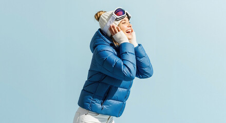 A happy young woman in winter ski attire smiles joyfully. Cheerful person in a blue puffer jacket and goggles on a blue background with copy space