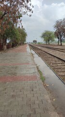Rural train station platform with wet bricks and parallel tracks under a cloudy sky, evoking a sense of tranquil travel and waiting.
