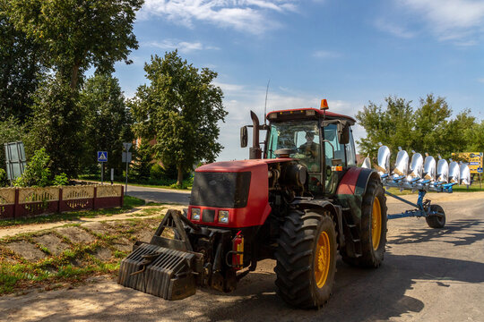 Agricultural tractor towing an 8-furrow plough in transport position on country road, showing curved moldboards for soil cultivation. Rural Belarus scene.