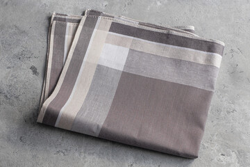 A single plaid handkerchief in a neutral palette of brown, beige, and gray is folded and displayed on a textured gray background.