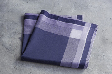 A folded men's handkerchief in shades of deep blue and purple with a classic plaid design is positioned on a rustic gray surface.
