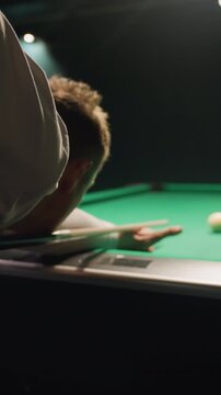 Close-up backshot of player in white shirt leaning over green pool table, gripping cue stick with intense focus. Dark background enhances concentration as he prepares to strike white cue ball