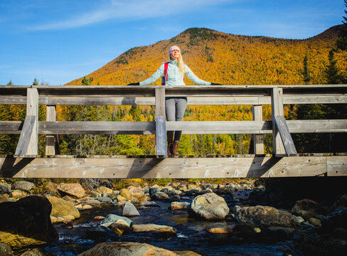 Hiker stands on wooden bridge surrounded by autumn foliage in mountain landscape during sunny afternoon