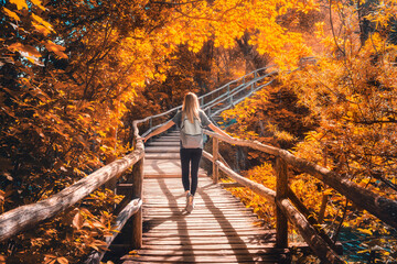 Young woman with backpack walking along a wooden path near stunning waterfalls, orange trees in Plitvice Lakes national park, Croatia at sunset in autumn. Sporty girl in forest in fall. Hiking, travel