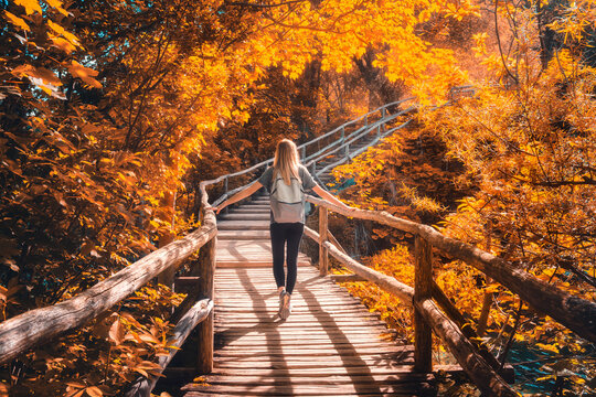 Young woman with backpack walking along a wooden path near stunning waterfalls, orange trees in Plitvice Lakes national park, Croatia at sunset in autumn. Sporty girl in forest in fall. Hiking, travel