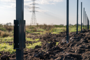 Steel fence posts aligned in soil at a construction site, with metal brackets attached, in a rural area with power lines in the background.