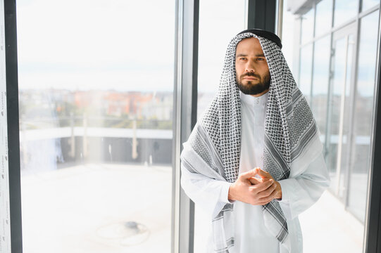 Arab businessman wearing traditional kandura standing next to office window - Powered by Adobe