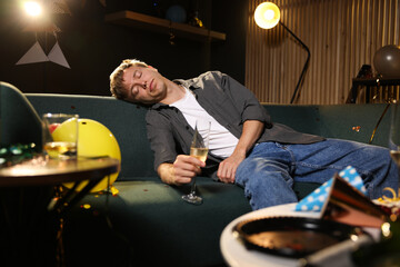 Man with glass of alcohol suffering from hangover on sofa in messy room after party