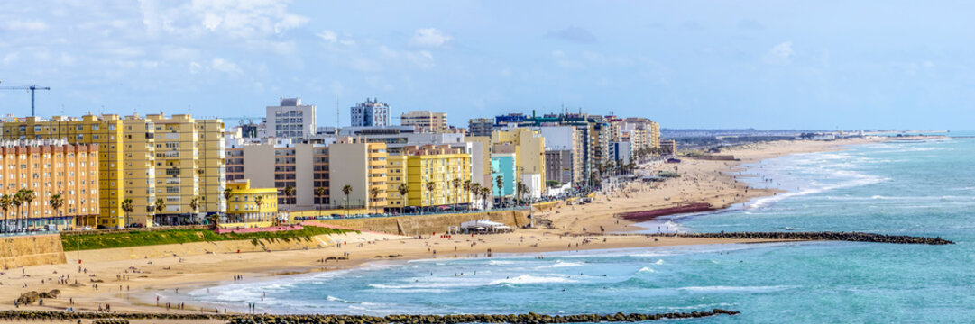 Wide panoramic view of the popular Playa de la Victoria in C&aacute;diz, with its calm waters and golden sand in the sunlight, while the city buildings rise on the horizon