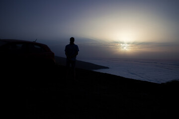 Silhouette of a person standing by a car, admiring a stunning sunset over a sea of clouds from a mountain viewpoint.