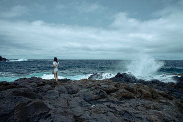 Woman standing on volcanic rocks near crashing ocean waves. Overcast sky and wild seascape create a dramatic coastal scene.