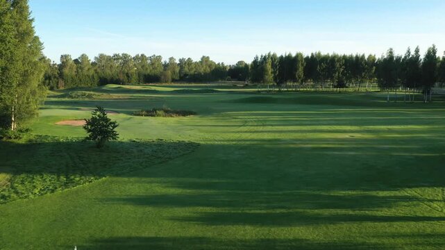 Morning sun casts long shadows on Avoti Golf Course, showcasing manicured greens, bunkers, and fairways surrounded by forest in a peaceful countryside setting.
