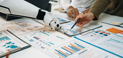 A robot hand helps a diverse team analyze business data in a modern office setting, showcasing charts and graphs essential for strategic decision-making.Noogenesis