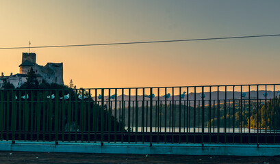 A row of seagulls perches on a railing with a medieval castle in the background at sunset. The warm tones of the sky contrast with the cool shadows, creating a scene of quiet balance between nature an
