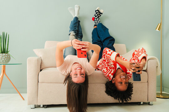 Two joyful females lying upside down on a couch, each engrossed in their mobile phones as they share a delightful experience using various apps and enjoying their time together in a lively and - Powered by Adobe