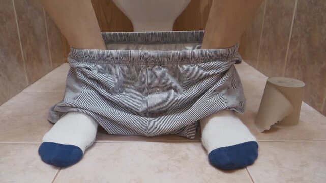 Man sits on toilet sink in restroom and impatiently twitches his legs. Man pushing, can not poop or suffer from constipation. Feet of man sitting on toilet in white socks and with his underwear down