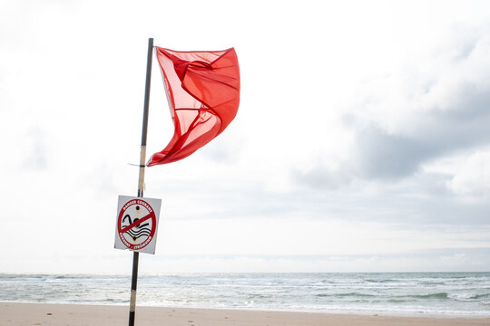 Red warning flag with danger current sign on sandy beach by the ocean, symbolizing hazardous swimming conditions and safety alert under cloudy sky.
