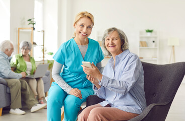 Portrait of a happy woman caregiver assisting an elderly patient with online technology using a phone in a nursing home. Supportive role of nurses and caregivers in modern elder care.