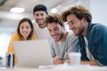 Obraz premium A group of smiling young professionals collaborates on a laptop. Represents teamwork, innovation, and a positive work environment. Ideal for illustrating startups, creative agencies, or education.