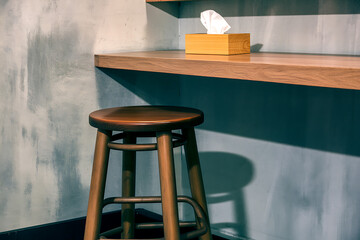 A simple wooden bar stool stands empty next to a concrete wall with a wooden shelf holding a tissue box